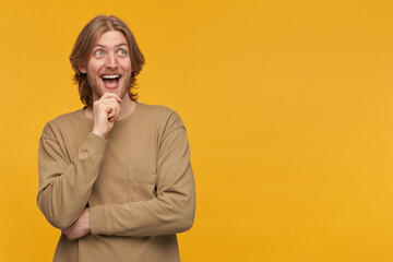 Cheerful male, positive and excited bearded guy with blond hairstyle. Wearing beige sweater. Touching his chin and smiling. Watching to the right at copy space, isolated over yellow background