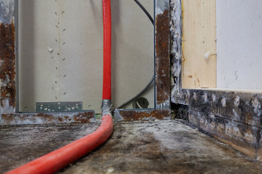 Drywall (Trockenbau) with rusty metal rails and red isolated water pipe. Mold and water stains on brown wood floor. Insurance claim and construction drying.