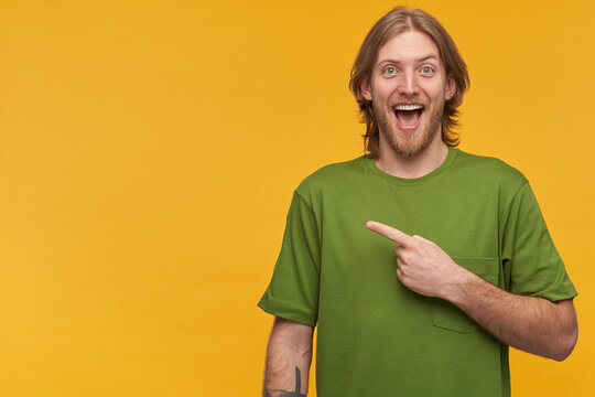Happy male, handsome bearded guy with blond hairstyle. Wearing green t-shirt. Has tattoos. Watching at the camera and pointing finger to the left at copy space, isolated over yellow background