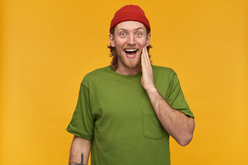 Young man, happy guy with blond hair, beard and mustache. Wearing green t-shirt and red beanie. Has tattoo. Touching his cheek. Watching at the camera isolated over yellow background