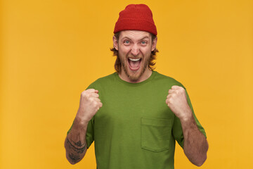 Young man, excited guy with blond hair, beard and mustache. Wearing green t-shirt and red beanie. Has tattoo. Clench his fists. Watching at the camera isolated over yellow background