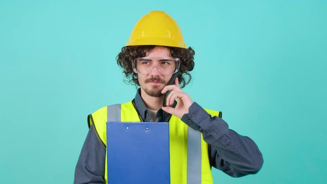 Handsome young construction worker with safety glasses and helmet that is thinking while holding clipboard and talking on mobile phone against turquoise background.
