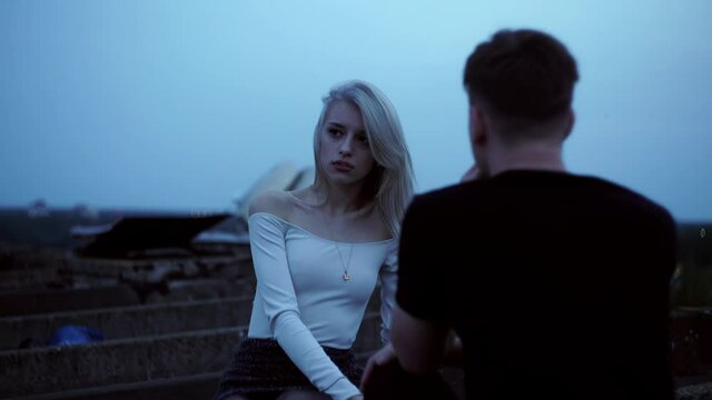 A very beautiful stylish girl is sitting on the roof next to her boyfriend and smoking. It's evening outside.