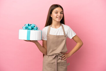Little girl with a big cake over isolated pink background posing with arms at hip and smiling