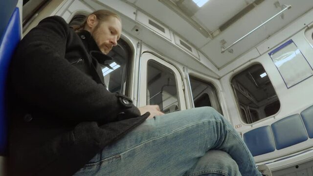 White man with beard and long blond hair sits in moving and shaking subway carriage. Guy scrolls through his phone