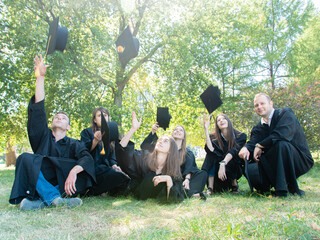 A group of young graduate students in gowns sitting in the park on the lawn cheerfully toss an...