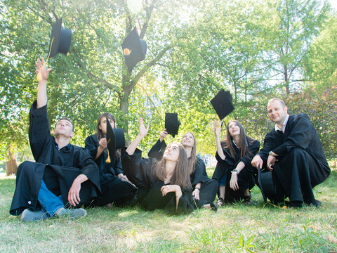 A group of young graduate students in gowns sitting in the park on the lawn cheerfully toss an academic cap up into the air