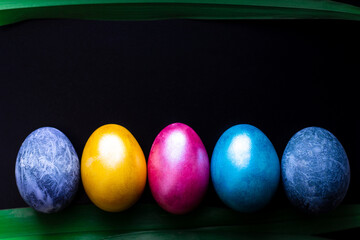 Multi-colored painted Easter eggs are lined up in a row on a black background with a dark green leaf at the bottom and a place to write in the center