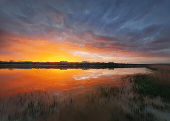 Fascinating sunset reflecting on the lake surface. Idyllic landscape, vertical background. Silent and tranquil evening scene with colorful sky, reed vegetation in the pond and a city at the horizon