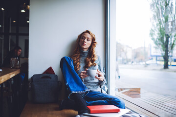 Young thoughtful woman having coffee sitting on window sill in cafeteria