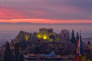 Illuninated Castle Ruin Baden with the roof of the St. Paul Church, colord by the twilight sky, in Badenweiler a historical town in Baden-Wuerttemberg, South Germany
