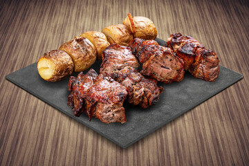 A selection of various barbecued gourmet meats on a black board with a rustic timber background. Pork neck, potatoes, kebab