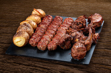 A selection of various barbecued gourmet meats on a black board with a rustic timber background. Rack of lamb, potatoes, kebab