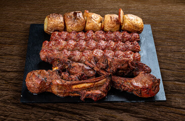 A selection of various barbecued gourmet meats on a black board with a rustic timber background. Rack of lamb, potatoes, kebab