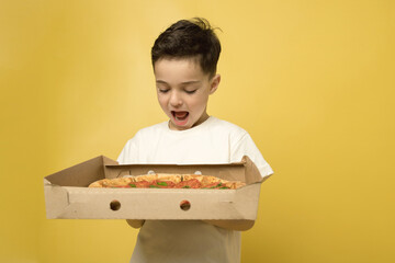 Brunet child posing with pizza in delivery box isolated on yellow background. Funny hungry boy is holding big pizza in a to go box. Caucasian. Fast food, junk food, children love pizza concept. 