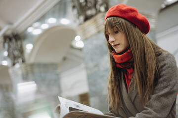 Beautiful Caucasian woman wearing warm coat, beret and scarf sitting alone on Saint Petersburg metro station reding a magazine. Image with selective focus