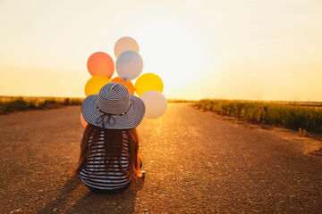 Little girl sitting on road with colorful balloons in hands at sunset. Back view