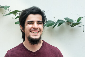 Portrait of young caucasian black haired man with beard and red shirt looking at camera, laughing and smiling naturally, confident and friendly. White background and climbing plant with leaves behind