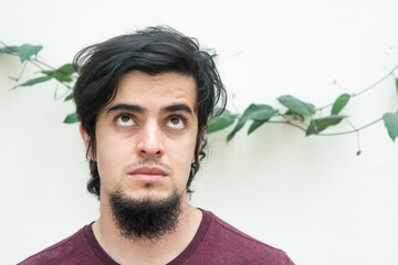Portrait of young caucasian black haired man with beard and red shirt looking up with doubt and fun expression naturally, confident and friendly. White background and climbing plant with leaves behind