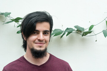 Portrait of a young caucasian black haired man with beard and red shirt looking at camera and smiling naturally, confident and friendly. White background and climbing plant with leaves behind