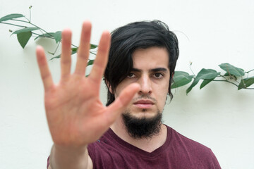 Young caucasian black haired man with beard and red shirt looking at camera with his hand on the front
making a stop hand sign. White background and climbing plant with leaves behind