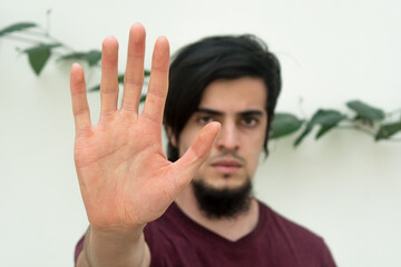Young caucasian black haired man with beard and red shirt looking at camera with his hand on the front
making a stop hand sign. White background and climbing plant with leaves behind