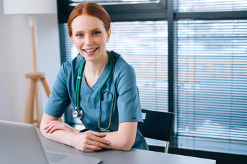Portrait of attractive smiling young female doctor in blue green medical uniform sitting at desk...