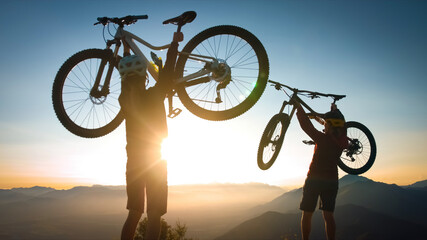 Two cheerful woman lifts his bicycle above his head at sunset after a winning mountain biking ride.