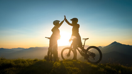 Two happy woman high five over the sunset after a successful mountain biking trip in the mountains....