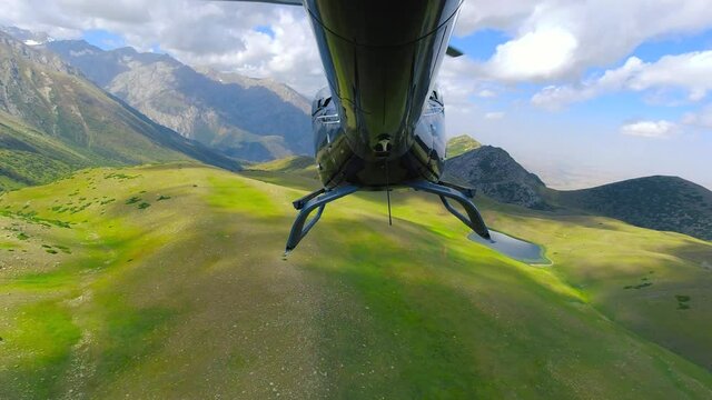 Helicopter view from the tail section flying over rocky mountains and green meadows.