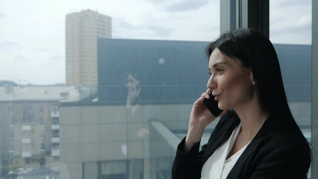 Portrait of happy young woman standing in front of office window having a phone conversation. Entrepreneur in a good mood, smiling, discussing news or success at work. Concept of technology, working