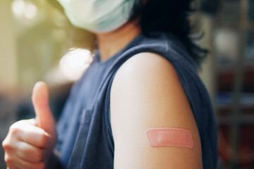 Close-up adhesive bandage on unrecognized person's arm after injection of vaccine, people in face mask received a coronavirus COVID-19 vaccine and giving thumb up to recommended inoculation vaccine. - obrazy, fototapety, plakaty