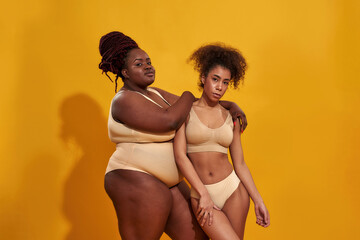 Two serious african american women in beige underwear with different body size standing together,...