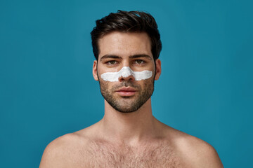 Portrait of handsome naked man with white mask applied on nose for cleansing pores posing isolated over blue background. Skincare routine concept