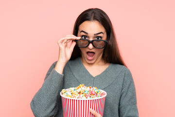 Young caucasian woman isolated on pink background surprised with 3d glasses and holding a big bucket of popcorns