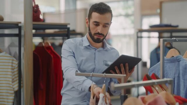 Clothing Store: Male Visual Merchandising Professional Uses Tablet Computer To Create Collection. Fashionable Shop Sales Retail Manager Checks Stock. Big Mall SUpermarket with Stylish Merchandise