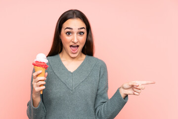 Young woman holding a cornet ice cream isolated on pink background surprised and pointing finger to the side