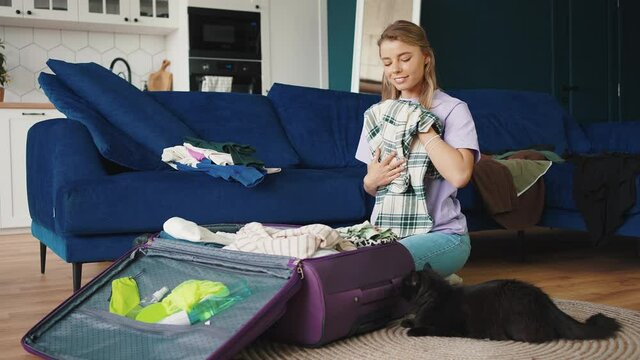 Young positive blonde woman packing clothes into suitcase and closing it, smiling to her lying on floor cat