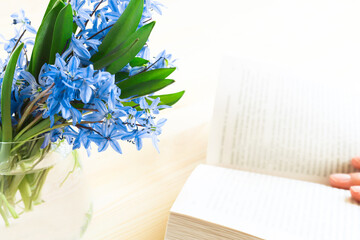 Open book with text on a wooden background. Blue flowers with green leaves in a glass vase in the foreground in focus. Vintage style and retro.