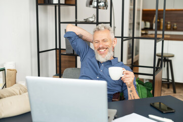 Fototapeta premium Cheerful mature male employee enjoys morning coffee at the workplace in the office, sits in relaxed pose with feet on the table, satisfied middle aged man in smart casual wear takes a break