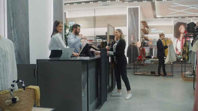 Clothing Store Checkout Cashier Counter: Beautiful Young Woman Buys Blouse from Friendly Retail Sales assistant, Paying with Contactless Credit Card. Trendy Fashion Shop with of Designer Brands
