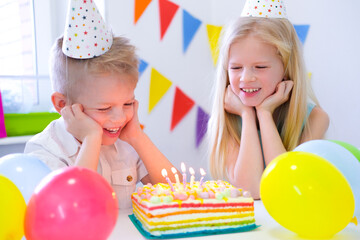 Fototapeta na wymiar Two blonde caucasian kids boy and girl have fun blowing out candles at birthday rainbow cake with burning candles at birthday party