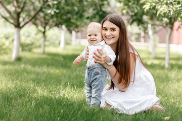 Happy beautiful woman, young mother playing with her adorable baby son, cute little boy, enjoying together a sunny warm day playing on the lawn in a summer garden