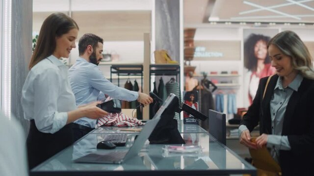 Clothing Store Checkout Cashier Counter: Beautiful Young Woman Buys Clothes from Friendly Retail Sales assistant, Paying with Contactless Credit Card. Fashion Shop with of Designer Brands. Medium Shot