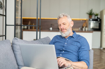 Middle-aged bearded man lying down on the couch with a laptop, working remotely from home, senior charismatic male in casual shirt typing, sending emails, messaging in relaxed home atmosphere