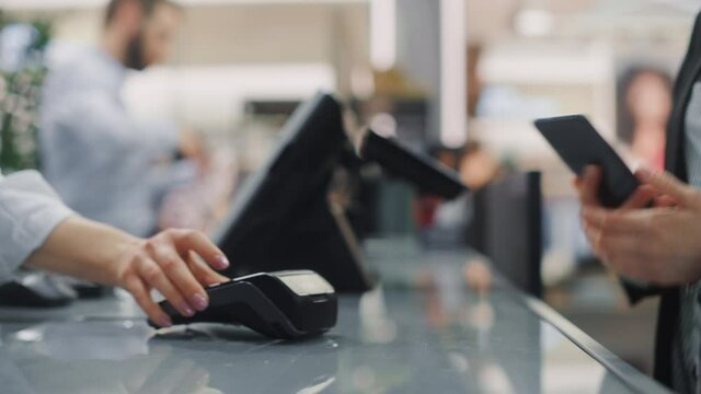 Clothing Store: Woman At Counter Buys Clothes Paying with Smartphone Through, Contactless NFC Terminal. Trendy Fashion Shop with Stylish Designer Brands. Close-up Focus on Hand