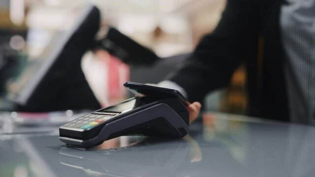 Clothing Store: Woman At Counter Buys Clothes Paying with Smartphone Through, Contactless NFC Terminal. Trendy Fashion Shop with Stylish Designer Brands. Close-up Focus on Hand