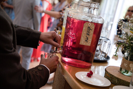 Mans hands pouring a blackcurrant drink or cocktail from a kilner jar that says drink me