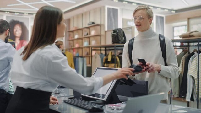 Clothing Store: Young Man At Checkout Counter Buys Clothes Paying with Contactless NFC Smartphone, Friendly Retail Sales Assistan Packs Merchandise. Contemporary Fashion Shop with of Designer Brand