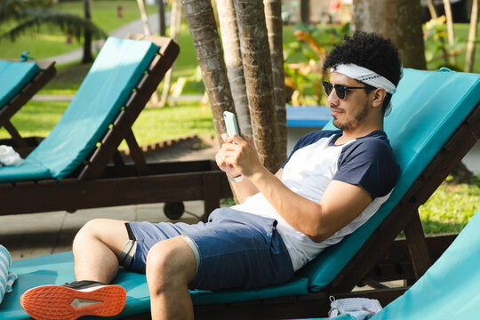 Hispanic young man sitting in a long chair by the pool while using his phone - Young man on vacation using his cell phone